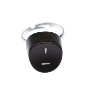 Puede incluir: Un anillo negro con una banda plateada y un signo de exclamación blanco y un guion en la parte superior.