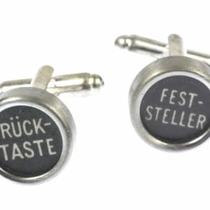 May include: A pair of silver cufflinks with black typewriter keys. The left cufflink says "RÜCK-TASTE" and the right cufflink says "FEST-STELLER".