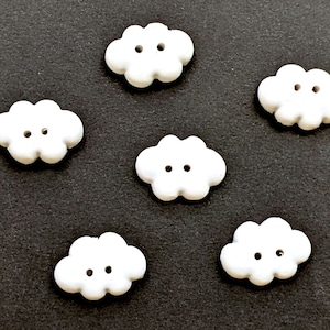 Puede incluir: Seis botones blancos en forma de nube con dos agujeros cada uno. Los botones están dispuestos en semicírculo sobre un fondo negro.