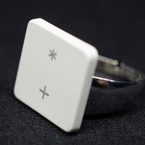 May include: A white ring with a square top featuring a plus sign and an asterisk. The ring is made of metal and has a silver finish.