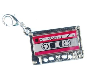 Puede incluir: Un charm de cinta de cassette plateado con un cierre de mosquetón. La cinta de cassette tiene un diseño rojo y blanco con la palabra "LOVE" y detalles de estrellas. Un accesorio nostálgico.