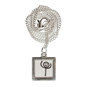 Può includere: Una collana con catena d'argento con un piccolo ciondolo a forma di cornice d'argento. La cornice contiene un disegno in bianco e nero di un omino stilizzato con un viso sorridente.