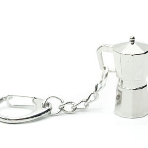 Pode incluir: Um chaveiro de metal prateado com um pequeno amuleto de cafeteira de metal prateado. O chaveiro tem um anel de metal prateado e uma corrente de metal prateado.