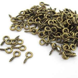 Può includere: Un mucchio di piccoli occhielli a vite color ottone antico su sfondo bianco. Gli occhielli hanno un'estremità a vite e una parte superiore ad anello, ideali per vari progetti di artigianato e gioielleria. Gli occhielli sono lunghi circa 1,3 cm.