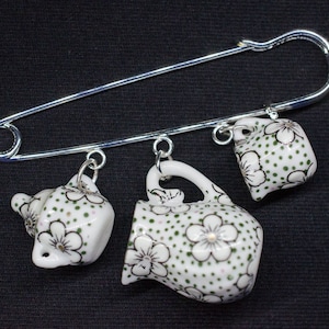 Puede incluir: Un broche de seguridad plateado con cuatro teteras de cerámica en miniatura colgando de él. Las teteras son blancas con un patrón floral verde y negro.