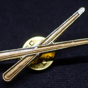 May include: A gold-toned pin featuring a crossed pair of drumsticks. The drumsticks have a detailed wood grain appearance and white tips. The pin is set on a gold-colored circular base, perfect for a drummer or music enthusiast.