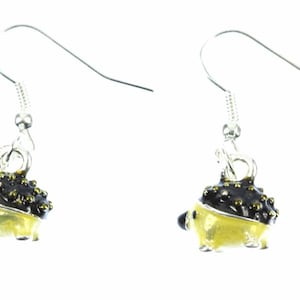 Peut inclure: Une paire de boucles d'oreilles pendantes argentées avec un petit charme de hérisson détaillé. Le hérisson est peint en jaune avec des accents noirs et dorés.