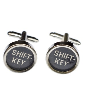 May include: A pair of silver cufflinks with black round buttons that say "SHIFT-KEY" in white lettering.