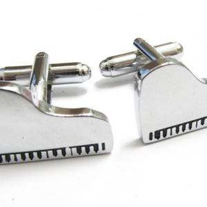 May include: A pair of silver-toned cufflinks shaped like grand pianos. Each cufflink features a piano keyboard design with black keys. The cufflinks have a shiny, reflective surface and a classic bar closure.
