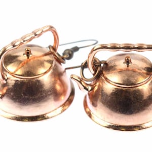 Peut inclure: Une paire de boucles d'oreilles en forme de théière de couleur cuivre. Les théières ont un corps rond, un couvercle, un bec verseur et une poignée. Les boucles d'oreilles sont attachées à un crochet de couleur argent.