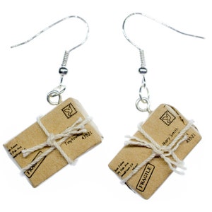 Peut inclure: Une paire de boucles d'oreilles en forme de colis en papier brun avec des crochets argentés. Les colis sont attachés avec de la ficelle et portent les mots "Fragile" et "Mary Smith" imprimés dessus.