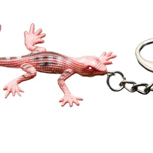 Peut inclure: Un porte-clés lézard rose avec un corps texturé et écailleux présentant des taches gris foncé sur le dos. Le lézard est fixé à un anneau fendu en métal argenté par une courte chaîne, formant un accessoire fonctionnel pour clés ou sacs.