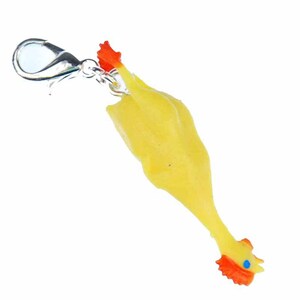 Könnte beinhalten: Ein gelber Gummi-Hühner-Anhänger mit orangefarbenem Kamm und Kehllappen sowie blauen Augen. Der Anhänger ist an einem silberfarbenen Karabinerverschluss befestigt. Dieser Artikel ist ein lustiges Accessoire.