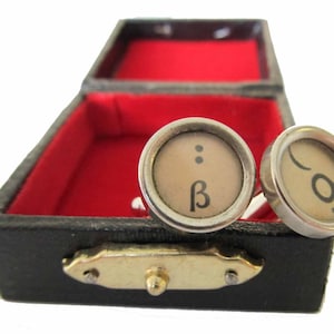 May include: Silver-toned cufflinks featuring typewriter keys, one with the Greek letter beta and dots, the other with the number 6. Presented in a black box with a red velvet lining and a gold-coloured clasp.