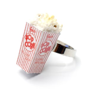 Könnte beinhalten: Ein silberner Ring mit einem Popcorn-Eimer-Anhänger. Der Eimer ist rot und weiß mit den Worten "Fresh Pop Corn" und "Delicious Crisp" bedruckt. Das Popcorn ist weiß und fluffig.