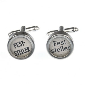May include: A pair of silver cufflinks with a round, white background and black text that reads "FEST-STELLER".