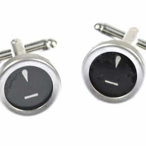 May include: A pair of silver cufflinks with black circular faces. Each face has a white exclamation point and a white horizontal line.