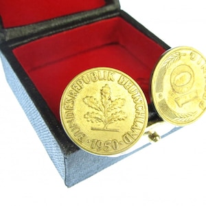 May include: A pair of gold cufflinks in a red velvet box. The cufflinks are shaped like German 10 Pfennig coins, one with the text "Bundesrepublik Deutschland 1950" and an oak leaf design, and the other with the number "10" and the text "Pfennig".