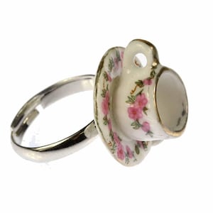 Könnte beinhalten: Ein silberner Ring mit einem daran befestigten Miniatur-Porzellan-Teetassen-Set. Die Teetasse und die Untertasse sind mit rosa Blumen und goldenen Verzierungen verziert.