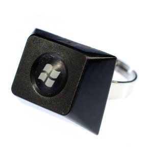 Könnte beinhalten: Ein schwarzer Ring mit einer quadratischen Oberseite, auf der das Windows-Logo abgebildet ist. Der Ring ist aus Metall und hat ein silbernes Band.