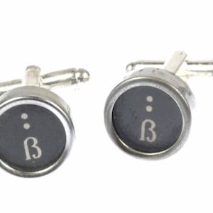 May include: A pair of silver cufflinks with black and white typewriter keys. The keys feature the letter "B" and two dots.