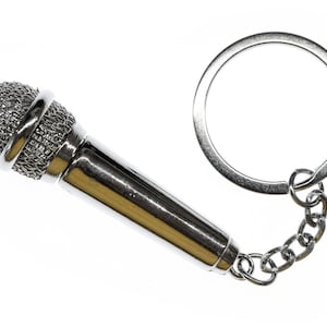 Könnte beinhalten: Ein Miniatur-Schlüsselanhänger in Form eines silbernen Mikrofons. Der Mikrofonkopf ist ein silbernes Netz, und der Griff ist glänzend silberfarben. Eine kleine Kette verbindet das Mikrofon mit einem silbernen Schlüsselring.