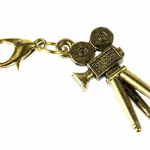 Könnte beinhalten: Ein goldfarbener Anhänger mit einem Vintage-Filmkamera-Design. Der Anhänger hat einen detaillierten Kamerakörper, zwei Filmrollen und eine Stativbasis. Er enthält einen Karabinerverschluss zur einfachen Befestigung an einem Armband oder einer Halskette.