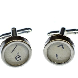 May include: A pair of silver cufflinks featuring typewriter keys. The keys are white with black lettering. The left cufflink has the letter 'é' and a slash. The right cufflink has a comma and a backwards apostrophe.