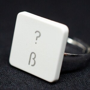 May include: A white ring with a square top that has a question mark and the letter B on it.