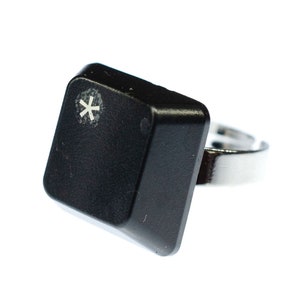 May include: A black computer key shaped ring with a silver band. The key has a white asterisk symbol on it.