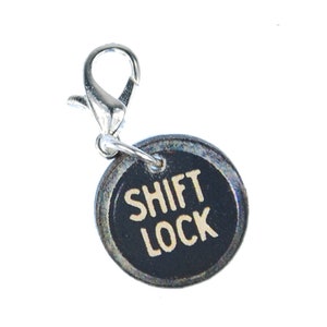 May include: A silver charm with a black round disc that says "SHIFT LOCK" in white letters. The charm has a silver lobster clasp.