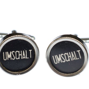 May include: A pair of silver cufflinks with black round buttons that say "UMSCHALT" in white lettering.