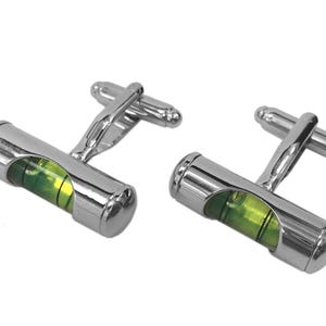 Può includere: Una coppia di gemelli in metallo color argento dal design cilindrico, ciascuno dotato di una fiala a bolla d'aria con liquido verde. I gemelli presentano una finitura metallica lucida e un meccanismo di chiusura a levetta standard.