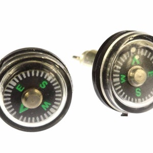 Puede incluir: Un par de pendientes de brújula negros con un borde plateado y una cubierta de plástico transparente. La cara de la brújula es negra con marcas blancas y una flecha verde que apunta al norte. Las letras N, S, E y W están impresas en verde.