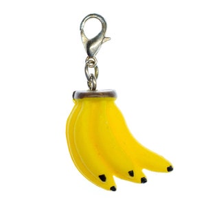 Op de afbeelding: Een gele banaanbedel met een zilveren clip. De bedel is gevormd als een tros bananen met zwarte puntjes.