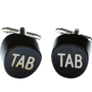 Peut inclure: Une paire de boutons de manchette noirs avec des accents argentés. Chaque bouton de manchette présente un bouton noir avec le mot "TAB" en lettres blanches.