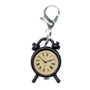 May include: A miniature black alarm clock charm featuring a cream-colored clock face with black Roman numerals and thin black hands. The charm is attached to a silver-toned metal lobster claw clasp, designed for easy attachment to various accessories.