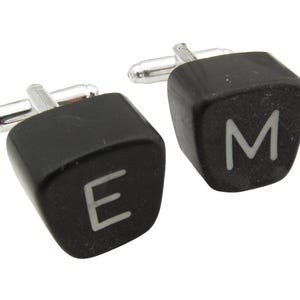 May include: A pair of black cufflinks shaped like typewriter keys. The keys have the letters "E" and "M" on them.