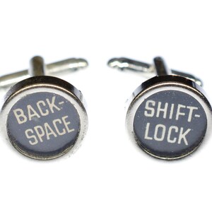Könnte beinhalten: Ein Paar silberne Manschettenknöpfe mit schwarzen runden Knöpfen. Die Knöpfe haben weißen Text, der "BACK-SPACE" und "SHIFT-LOCK" lautet.