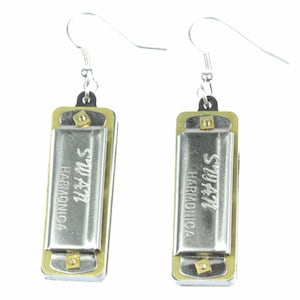 May include: A pair of silver harmonica earrings. Each earring features a small, silver harmonica with the words "SWATI HARMONICA" engraved on the front. The harmonicas are attached to silver hooks.