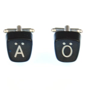 May include: A pair of black cufflinks shaped like typewriter keys. The left cufflink has the letter "A" with two dots above it. The right cufflink has the letter "O" with two dots above it.