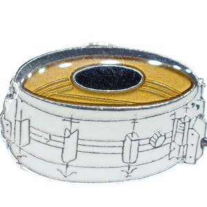 May include: A white and silver snare drum pin with a gold-colored drumhead and a black oval center. The pin features detailed outlines of the drum's hardware and is a musical accessory.