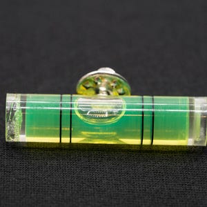 Op de afbeelding: Een klein, groen en geel waterpas met een metalen bevestigingsbeugel. Het waterpas is met een schroef aan een oppervlak bevestigd.
