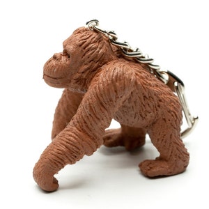 Peut inclure: Un porte-clés en forme d'orang-outan brun avec une chaîne argentée. L'orang-outan est fait d'un matériau texturé et a une apparence réaliste.