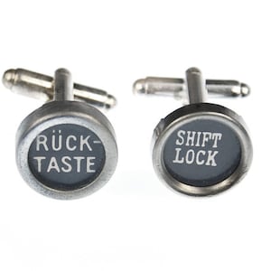 May include: A pair of silver cufflinks with black round buttons. The left cufflink has the text "RÜCK-TASTE" on the button. The right cufflink has the text "SHIFT LOCK" on the button.