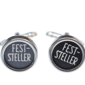 May include: A pair of silver cufflinks with black circular faces. Each face has the text "FEST-STELLER" printed in white.