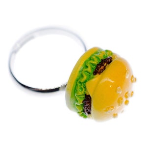 Könnte beinhalten: Ein silberner Ring mit einem gelben und grünen Hamburger-Anhänger. Der Hamburger-Anhänger hat ein Sesambrötchen, Salat und ein braunes Patty.