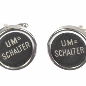 May include: A pair of silver cufflinks with black round buttons. The buttons have white text that reads "UM=SCHALTER".
