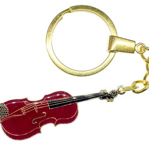 May include: A gold-toned keychain featuring a miniature violin charm. The violin is a deep red color with gold accents, including the strings and outline. The keychain has a gold ring and chain.
