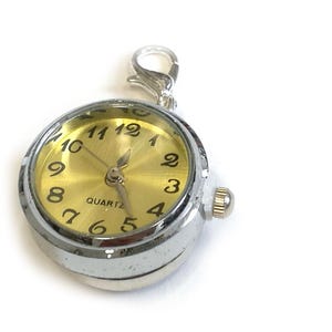 May include: A silver-toned quartz pocket watch with a gold-colored face. The watch has black numerals and hands, and a small crown on the side. It includes a silver clasp for attaching to a chain or bag.